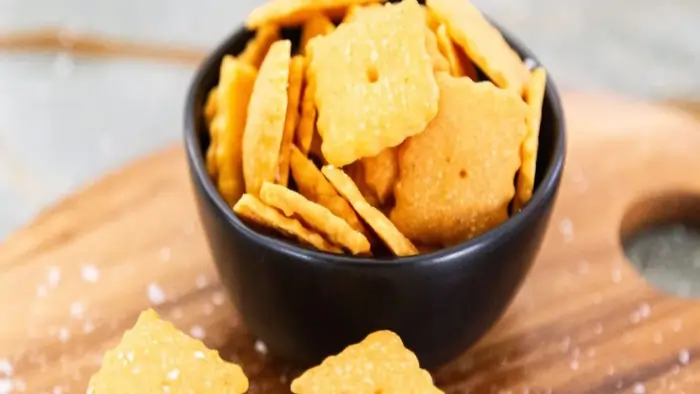 A black bowl filled with square, golden-brown gluten free cheez its, showing two additional crackers and sprinkled salt on a wooden board