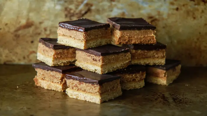 A stack of square-cut gluten free desserts featuring layers of shortbread, a creamy caramel or peanut butter center, and a smooth dark chocolate topping.