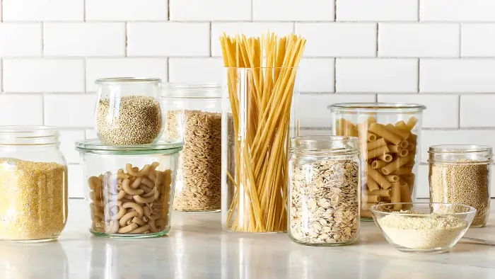 Gluten free grains and pasta displayed in glass jars including oats, quinoa, rice, spaghetti, macaroni, and flour on a kitchen counter.