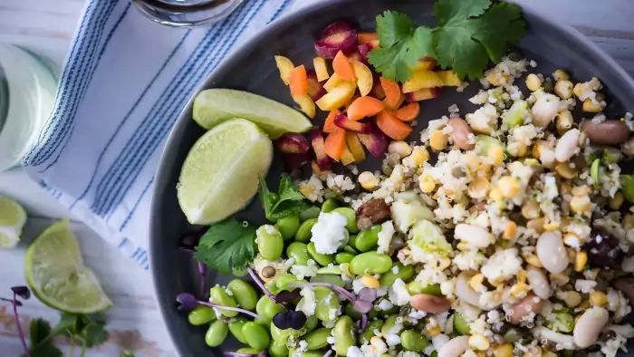 Healthy quinoa and bean salad bowl with Gluten-Free Whole Foods including edamame, mixed vegetables, lime wedges, and fresh cilantro on a table.